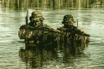 Pair of soldiers in action during river raid in the jungle waist deep in the water and mud and covering each other