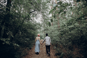 stylish hipster bride and groom walking in green summer forest. happy couple in love, modern outfit, relaxing at park. girl in dress and straw hat with peony. rustic wedding concept.