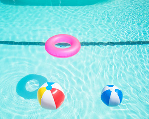 Bright pink float and beach balls in blue swimming pool, floating in refreshing swimming pool with waves reflecting in summer sun. Active vacation background. Lifesaver for kid. Sunny day at the pool