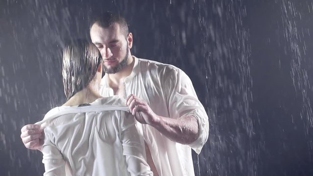 large man with a beard in a white shirt stands with his woman in the darkness under the shower. he takes off his shirt with shoulder wife Stripping naked slender back