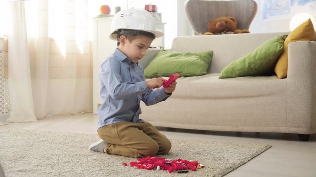 small kid boy imitates engineer constructing a building using colorful meccano details with enthusiasm and focus dreaming about future profession, sitting at home on floor
