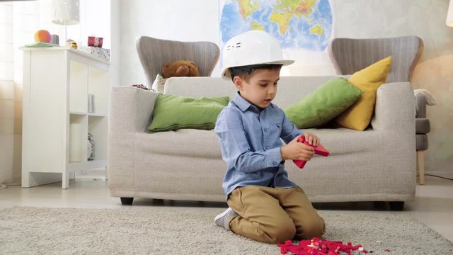 small kid boy imitates engineer constructing a building using colorful meccano details with enthusiasm and focus dreaming about future profession, sitting at home on floor