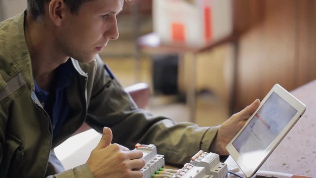 Electrician checks correct assembly of the switchboard on tablet. Young man with stubble on his face pokes a finger at the screen and said key n equipment.