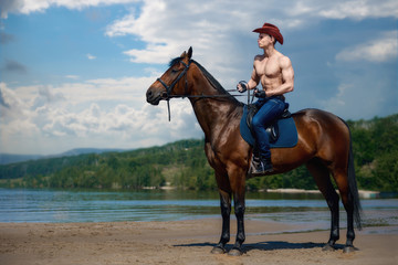 Macho man handsome cowboy riding on a horse on the background of sky and water.