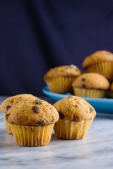 Three miniature vanilla chocolate chips muffins on marble surface in front of blue plate with some more muffins