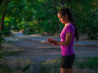 Shot of young caucasian woman standing in the forest with smart watch and put on it. Attractive teen tracking activity with fitness application.