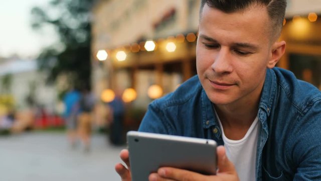 Portrait of handsome man in casual shirt using tablet while sitting on the bench in the city centre. Outdoor shooting.