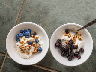 Two ceramic bowl with yogurt, crashed walnuts, blackberries and blueberries on a granite countertop