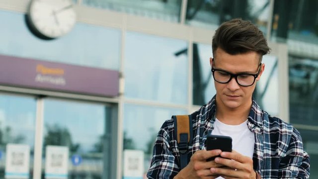 Young stylish man in glasses using smart phone for chatting in the network on the modern airport background. Close up.