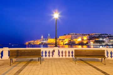 Summer night view of the embankment with benches and lantern and Valetta profile over sea at the background. Long exposure. Illuminated architecture.
