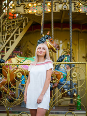 Young attractive blonde girl standing near carousel in amusement park. Beautiful portrait of woman with long hair.