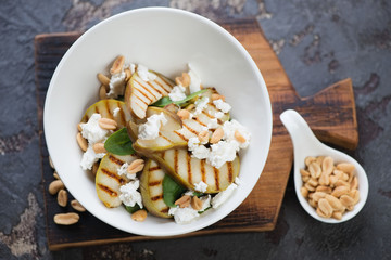 White bowl with grilled pears, spinach leaves, nuts and cheese salad on a wooden serving board, high angle view