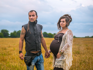 Man in a black cloth and pregnant woman in long dress with dreadlocks standing in wheat field on stormy sky background. Love story. Informal people with tattoo and piercing.