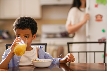little boy drinking orange juice at breakfast with mother in background