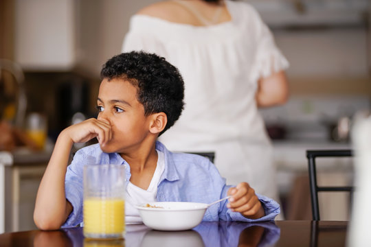 Little Mixed Race Boy Pondering While Eating Breakfast With Mother In Background