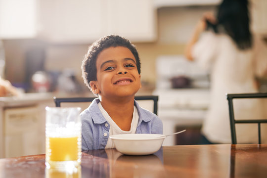 Cute Little Mixed Race Boy Smiling Into Camera With Mother In Background At Breakfast