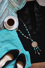 Stylish layout of clothes and accessories. Turquoise pants, evening top, black shoes, rosary and a cup of coffee