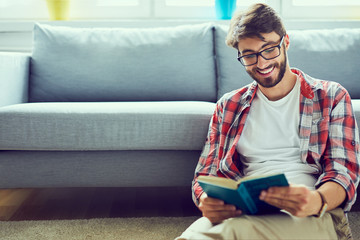Portrait of joyful young man reading book while sitting on floor in his living room - obrazy, fototapety, plakaty