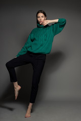Attractive Caucasian happy sexy female model with brunette hair is moving in studio, wearing green sweater, isolated on dark background
