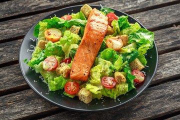 Caesar Salmon Salad with croutons, parmesan cheese, tomatoes, dressing and pepper in a black plate on wooden table.