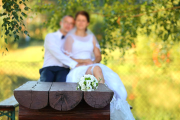 Bukiet ślubny na drewnianej ławie z młodą parą w tle. - obrazy, fototapety, plakaty
