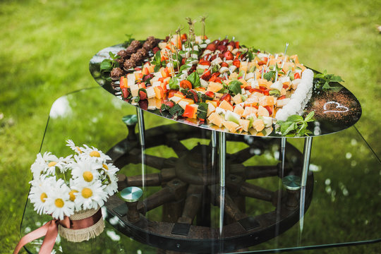 Catering for wedding party. Plenty of mixed various kinds of sliced fruit, vegetables,  cheese, chocolate sweets on big tray on table outdoors. Table decorated with vase full of white daisies flowers.