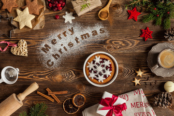 Words Merry Christmas on flour, wooden background. Christmass symbols sweets, candle, cinnamon, cup of coffee, dried orange, gift with red ribbon. View from above.