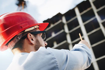 Young bearded engineer or architect in red hard hat with pencil in hand at construction site....