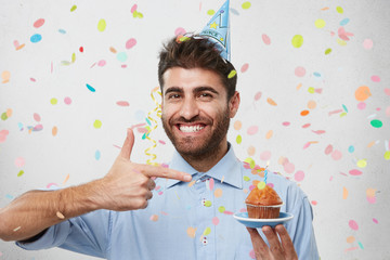 Happy bearded man with broad smile wearing party cap and formal shirt, holding little cake with...