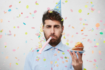 Displeased unhappy bearded Caucasian man with cone hat on head and party horn in mouth looking at...