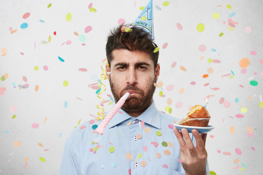 Displeased unhappy bearded Caucasian man with cone hat on head and party horn in mouth looking at camera with bored dissatisfied expression as his birthday party sucks. People and lifestyle