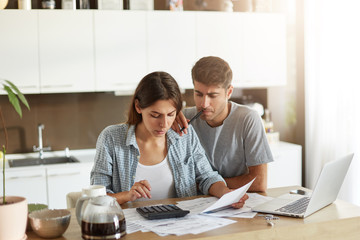 Young couple going to have wedding, calculating all their expenses, looking attentively at prices, planning what to order. Preoccupied man holding pen leaning at shoulder of her wife, looking in paper