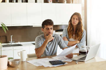 Frustrated young European couple facing financial crisis. Man with stubble sitting at kitchen table...