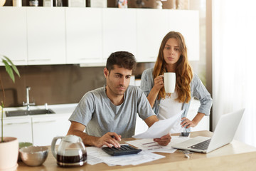 Candid shot of young American couple managing family budget, reviewing bank accounts using laptop and calculator in kitchen. Man and woman doing paperwork together, paying taxes online on notebook pc