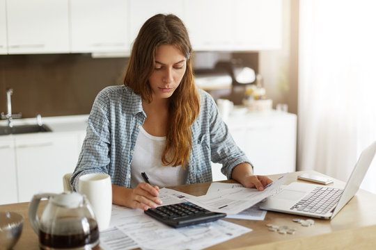 Picture of young Caucasian woman wearing casual clothes at home, full of sad and restless thoughts, feeling stressed calculating her debts for rent and making notes, surrounded with papers and gadgets