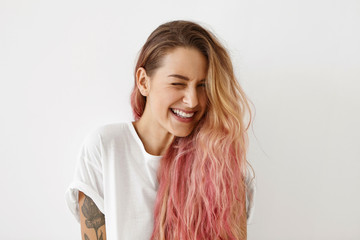 Young female model with pink hair tips, winking her eyes while having joy, smiling broadly, feeling...