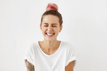 Gorgeous woman with pure healthy skin, pinkish hair knot and white perfect teeth closing her eyes with enjoyment being happy to find out that she is pregnant. Happy young woman laughing broadly - obrazy, fototapety, plakaty