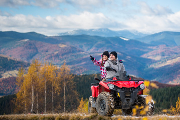 Young couple on red quad bike ride in mountains. Happy man driving the ATV, girlfriend pointing at something interesting. Beautiful landscape of mountains and forest at sunny day