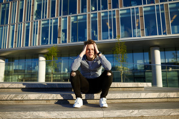 Handsome Caucasian guy wearing white sneakers, ripped jeans and shirt sitting on concrete stairs in urban setting, holding head, feeling unhappy and stressed out after big fight with his girlfriend