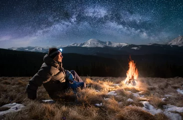 Fotobehang Male tourist have a rest in the mountains at night. Guy with a headlamp sitting near campfire under beautiful night sky full of stars and milky way, and enjoying night scene © anatoliy_gleb