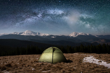 Night camping. Illuminated tourist tent under beautiful night sky full of stars and milky way. On the background snow-covered mountains and forests