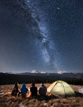 Rear view of four people sitting together beside camp and tent under beautiful night sky full of stars and milky way. On the background snow-covered mountains. Long exposure