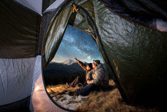 View from inside a tent on the two tourists have a rest in the camping in the mountains at night. Men sitting near campfire. One guy is pointing at the beautiful night sky full of stars and milky way
