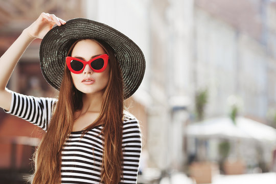 Outdoor portrait of young beautiful girl with long hair posing in street. Model wearing stylish red sunglasses, black hat, stripped blouse. City lifestyle. Sunny day light. Copy, empty space for text