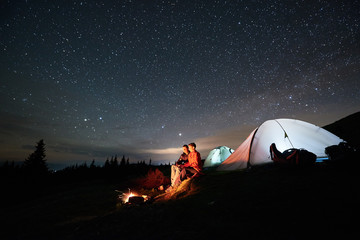 Night camping in the mountains. Man and woman tourists sitting at a campfire near two illuminated...