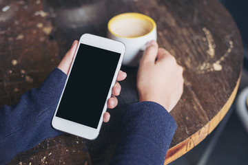 Mockup image of woman's hands holding white mobile phone with blank black screen and a coffee cup on wooden table in vintage cafe