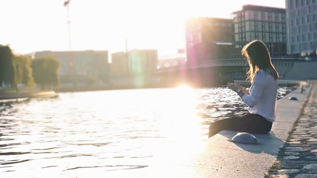 Beautiful girl taking a photo on smartphone sitting near the river at city center pedestrian in Berlin hd slow motion