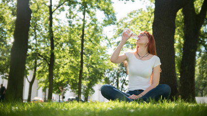 Naklejka na ściany i meble Woman in glasses sitting on grass in summer park and drinks water