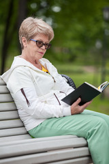 An elder woman in glasses reading black book while sitting on the bench in park