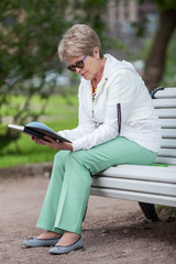 Fototapeta premium Full-length portrait of elderly woman reading book in hands, summer park